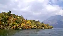 Photo couleur d'une pointe montagneuse et boisée, avancée dans une étendue d'eau (de gauche à droite). Un volcan (à droite) sous un ciel nuageux, en arrière-plan.