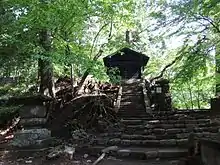 Photo couleur d'un édifice religieux en bois noir, au sommet d'un escalier en pierre et entouré d'arbres sous un ciel nuageux.