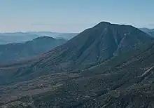 Photo couleur montrant une montagne aux pentes boisées et un massif montagneux au second plan, sous un ciel bleu.