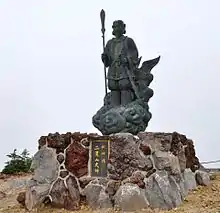 Photo couleur d'une statue en bronze posée sur un socle en pierre et représentant une divinité masculine sur un nuage, avec un ciel nuageux en arrière-plan.