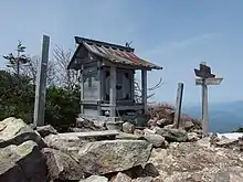 Photo couleur d'un sanctuaire shintō miniature en bois. Il y a un sol pierreux à l'avant-plan et un ciel bleu en arrière-plan.