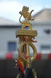Fleuron de hampe de drapeau belge avec couronne de feuilles de chêne et de laurier, la devise nationale et le lion héraldique.