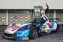 Photographie d'un homme assis sur le toit de sa voiture blanche et bleue, heureux et levant le poing.