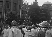 Photo noir et blanc datant de 1968 et montrant un groupe d'étudiants japonais, au premier plan et de dos. Ils sont tous casqués et armés d'une lance en bambou et ont le visage en partie masqué.