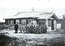 Photo d'un bâtiment en bois de plain-pied, devant lequel posent un groupe d'enfants et leur professeur.
