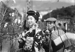 Une jeune femme (Kinuyo Tanaka) en kimino se tient debout près d'une barrière en bois. En retrait derrière elle, un homme en tenue de voyage tient d'une main une cigarette et de l'autre un bagage (image en noir et blanc).