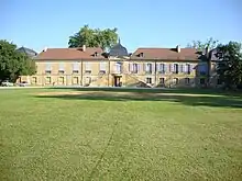 Un grand terrain vert avec à l'arrière-plan un long bâtiment à deux étages, de pierre ocre au toit de tuiles.