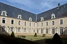 Photographie présentant un angle du jardin du cloître, dans un bâtiment à 2 niveaux de fenêtres et un toit en ardoise. Les fenêtres du rez-de-chaussée sont encadrées d'arcs surmontés d'angelots en clés de voûte. Au premier étage, sont visibles aux deux fenêtres de gauche deux mannequins en habits de religieuses.