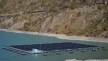 Au centre de la photo, on distingue des panneaux solaires flottant sur l'eau. En arrière plan, on aperçoit la paroi rocheuse plongeant dans le lac. On distingue une certaine hauteur de roche sans végétation indiquant que le lac de barrage n'est pas à son niveau le plus haut.