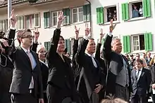 Photographie de quatre personnes, trois hommes et une femme, levant leur main droite au ciel avec le pouce, l'index et le majeur écarté.