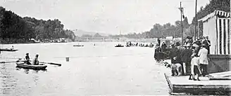 Photographie noir et blanc : des barques sur une large rivière ; une foule au bord de l'eau à droite.