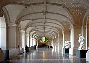 Vue du hall dans sa longueur, rythmé de lourd piliers de pierre sous une voute cintrée parsemée de lumignons, à droite des statues, à gauche des guichets, au fond un lustre multicolore.