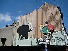 Photographie en couleur d'une fresque peinte sur le mur d'un bâtiment montrant trois personnages de cette série : un agent de police, avec un grand manteau noir, à gauche, et deux jeunes garçons à droite.