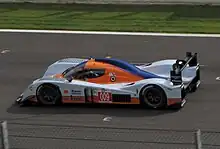 Photographie d'une voiture de sport-prototype, orange, bleu ciel et bleu foncée, de haut et de profil, sur une piste.