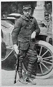 Photo d'un homme en uniforme tenant son arme et appuyé sur le passage de roue d'une voiture.