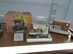 Deux machines à coudre dans une vitrine. Elles ont toutes les deux des coques en plastique clair. Celle de gauche, sans marque, a une teinte un peu argentée. Elle est exposée avec sa boîte d'origine, qui montre une photo de la machine sur un fond moitié rose, moitié jaune. À droite, la machine de marque Ma cousette est blanche, sauf la partie inférieure du bras et le centre de la tour qui sont bruns.
