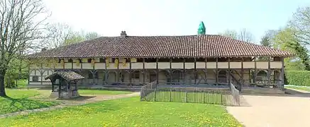 Un long bâtiment à colombages traversée sur sa longueur par un balcon et surmonté d'une cheminée sarrasine emballée dans un filet vert.