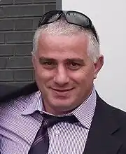 Photographie de la tête et du buste d'un homme aux cheveux gris, vêtu d'une chemise à rayures, d'une cravate et d'une veste noire