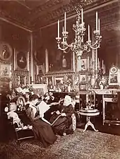 Photographie en noir et blanc d'une Victoria âgée assise à côté d'une jeune femme (Beatrice) lisant un journal. La pièce est richement décorée avec de nombreuses photographies et peintures et un large chandelier est suspendu au plafond.