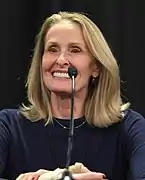 Photo en buste d'une femme âgée souriante avec des cheveux longs blonds et devant un micro.