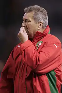 Un homme vu de profil et vêtu d'un k-way rouge, met ses doigts au-dessus de sa bouche.