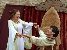 Photographie couleur d’un homme à genou tenant les mains d’une femme en blanc, debout et souriante.