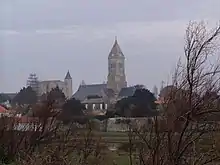 Photographie en couleur représentant un terrain vague dans le quartier de Banzeau, avec à l'horizon les tours de l'église Saint-Philbert et du château de Noirmoutier.