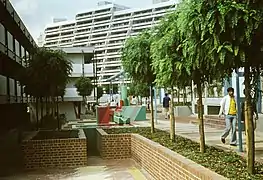 À l'avant-plan, une petite place avec un muret en pierre décorée. Quelqu'un se promène dans une allée bordée d'arbres. En fond, un grand immeuble et quelques canalisations surélevées.