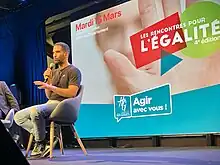 Ouissem parle dans un micro, assis sur une chaise sur un podium, avec l'affiche de l'évènement derrière lui