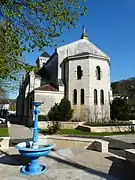 Photo représentant le chevet polygonal ensoleillé de l'église Saint-Jean-Saint-Charles surmonté d'une statue. À l'arrière, on peut voir une partie du clocher carré. Au premier plan sur la gauche se trouve une fontaine métallique de couleur bleu turquoise avec vasque.