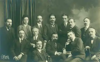 Photographie en noir et blanc de treize hommes. Six sont assis et les autres derrières sont debout. En arrière plan à gauche on voit un rideau qui cache en partie ce qui ressemble à un mur peint représentant un feuillage.