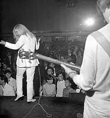 Photographie en noir et blanc, sur une scène, avec en premier plan, sur la droite partie dos tronqué sur la longueur d'une personne jouant de la guitare, au second plan, image féminine de dos, vêtue d'un costume blanc, jouant de la guitare, au fond, une foule de spectateurs