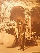 Un homme en costume sombre et chapeau dans un parc devant une loggia avec jarre.