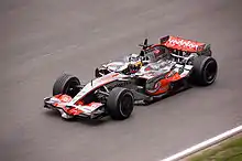 Voiture gris argenté aux bords rouges, vue de haut, de trois-quarts, sur une piste sèche.