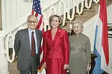 Ces trois personnes posent devant le drapeau américain et le drapeau du Luxembourg, le tout devant un escalier blanc.