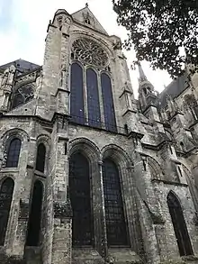 Petit transept, en partie caché par un arbre, avec sa rosace en forme de fleurs et les deux grandes verrières de la partie basse. À droite, bâtiment de brique.