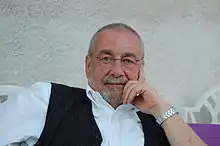 Portrait buste d'un homme assis grisonnant portant des lunettes, chemise et gilet dans une pause décontractée.