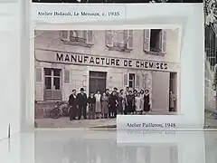 Photographie en noir et blanc. Devant un bâtiment à façade en pierre avec plusieurs fenêtres aux volets ouverts, au fronton duquel on lit « Manufacture de chemises », une quinzaine de personnes, dont une dizaine de femmes et 5-6 hommes, posent debout face à l'objectif. Tous et toutes ont des tenues sobres. Les femmes sont en robe longue ou pull et jupe longue et portent les cheveux courts ou en chignon. Les hommes sont en salopette et veste, ou en complet. Un homme sur la gauche porte un costume élégant avecu un chapeau sombre. Vers la droite, un autre porte un béret noir.