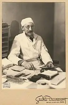 Photographie en tons sépias d'un homme assis, souriant, coiffé d'une toque blanche, tenant dans sa main droite un marteau à réflexes