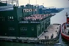 Un bâtiment au bout d'un quai, la façade marquée de l'inscription en grandes lettres « French Line ». Une foule attend sur la terrasse et salue l'arrivée du paquebot d'où la photo est prise.