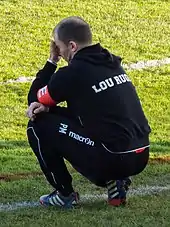 Un homme en survêtement sur le bord d'un terrain, accroupi.