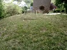 Vue en surplomb d'une terrasse engazonnée portant un cadre en bois et en contrebas, la base d'un monument.