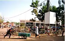 place en latérite avec quelques passants piétons, un arbre, un poteau électrique et une structure blanche sur le côté droit