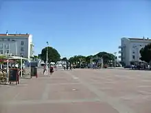 Photographie d'une vaste place vide au revêtement rosé quadrillé de pierres plates. Au fond la grille 1900 en fer forgé, les pins et palmiers du front de mer