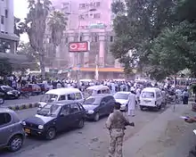 Photo montrant une scène de rue, au premier plan de nombreuses voitures circulent surveillées par un homme en armes, au fond des personnes sont rassemblées devant un immeuble