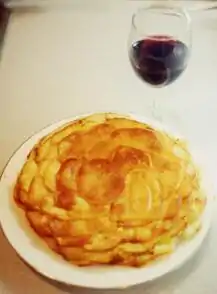 À côté d’un verre de vin rouge, un gâteau composé de fines tranches de pommes de terre superposées en dôme doré et croustillant est disposé sur une assiette blanche.