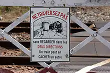 Gros plan sur le panneau émaillé du portillon qui porte l'indication : « NE TRAVERSEZ PAS sans regarder dans les DEUX DIRECTIONS. Un train peut en CACHER un autre » et un dessin avec une famille qui traverse alors que survient une locomotive à vapeur qui leur était masquée par un wagon.