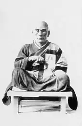 Photo noir et blanc d'une statue représentant un homme chauve, assis en tailleur sur un banc en bois et vêtu d'une robe traditionnelle japonaise (fond clair).