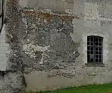 Photographie en couleurs d'un pan de mur au revêtement et parement en partie détruit sur la gauche, une fenêtre à carreau en plein-cintre à droite.