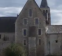 Photographie en couleurs d'un mur flanqué d'un contrefort, percé de sept fenêtres et terminé par une maçonnerie triangulaire, le tout encadré par des bâtiments à toits pentus, la flêche d'un clocher visible en arrière-plan.